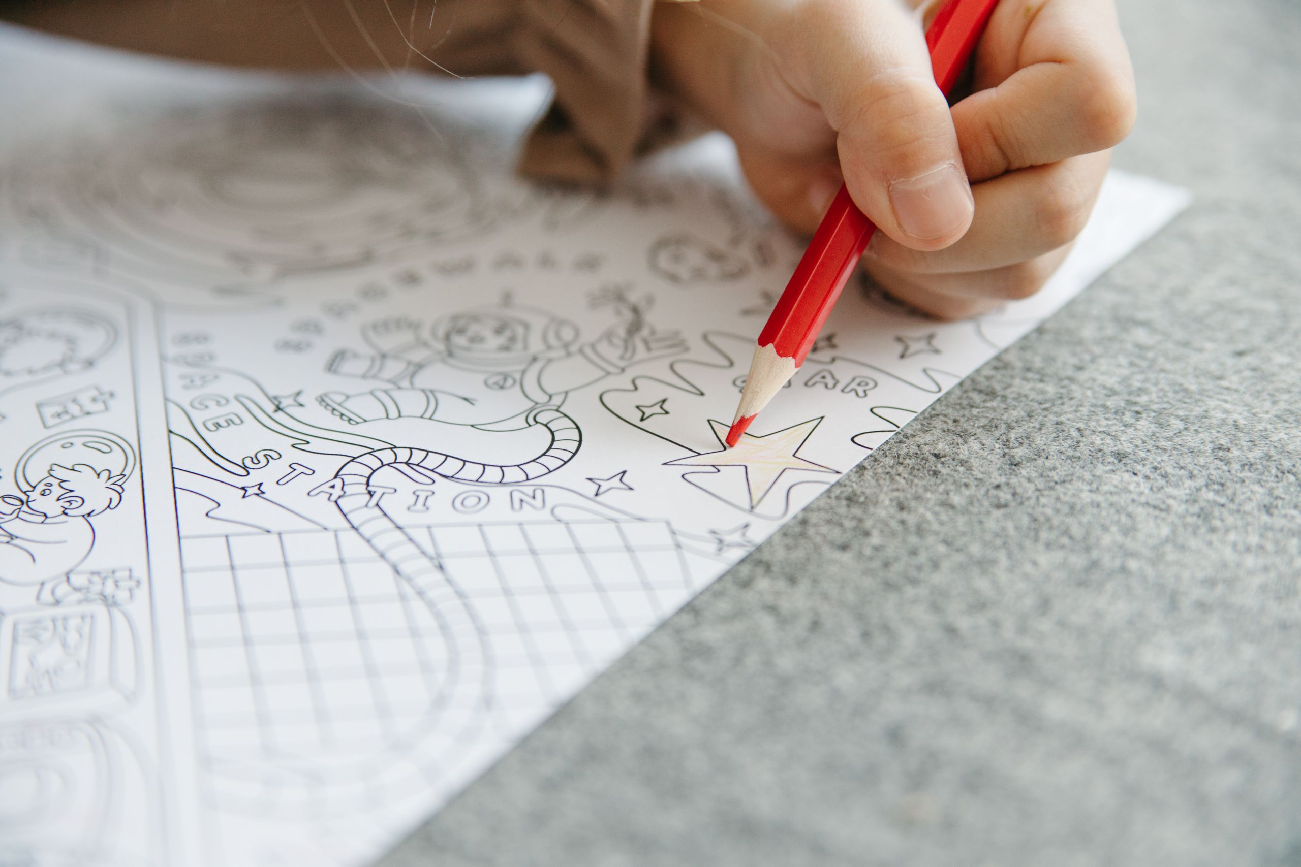 Close-up of a child’s hand using a red pencil to color a star in a space-themed drawing.